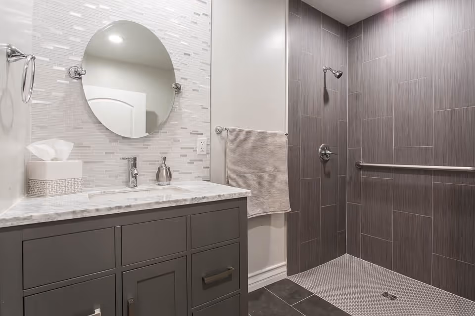 Modern bathroom with a gray vanity topped with a marble countertop, a round mirror above the sink, a tissue box, and a soap dispenser. The shower area features dark gray tiles, a wall-mounted showerhead, and a grab bar. A beige towel hangs on a towel rack next to the shower.