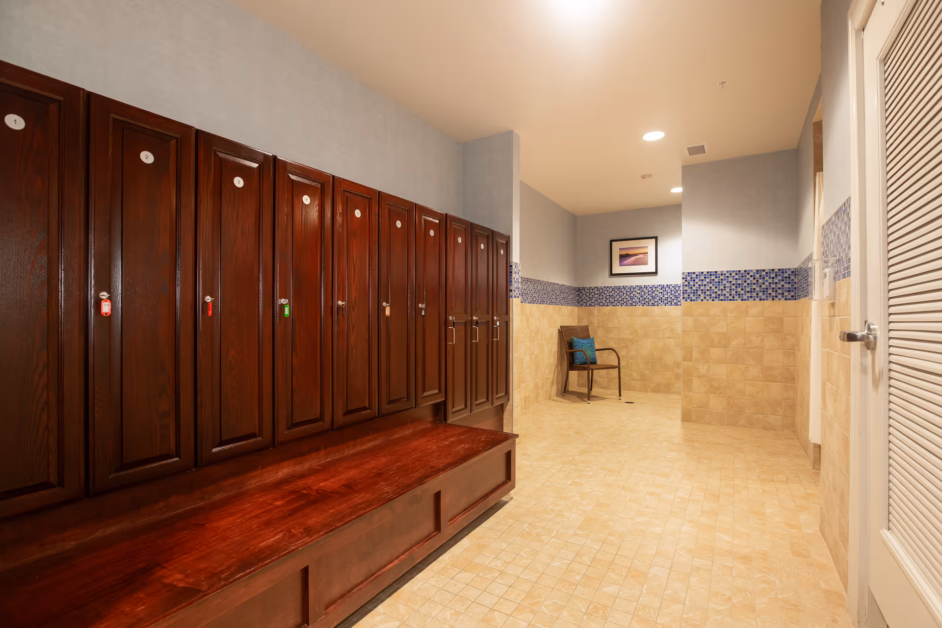 Interior view of a locker room with a row of dark wooden lockers on the left side, each with a numbered circular label and keychains. The floor is tiled in a light beige color, and the walls are partially tiled with beige tiles and a blue mosaic tile border. There is a single chair with a cushion against the far wall, and a framed picture hanging above it. The ceiling has recessed lighting.