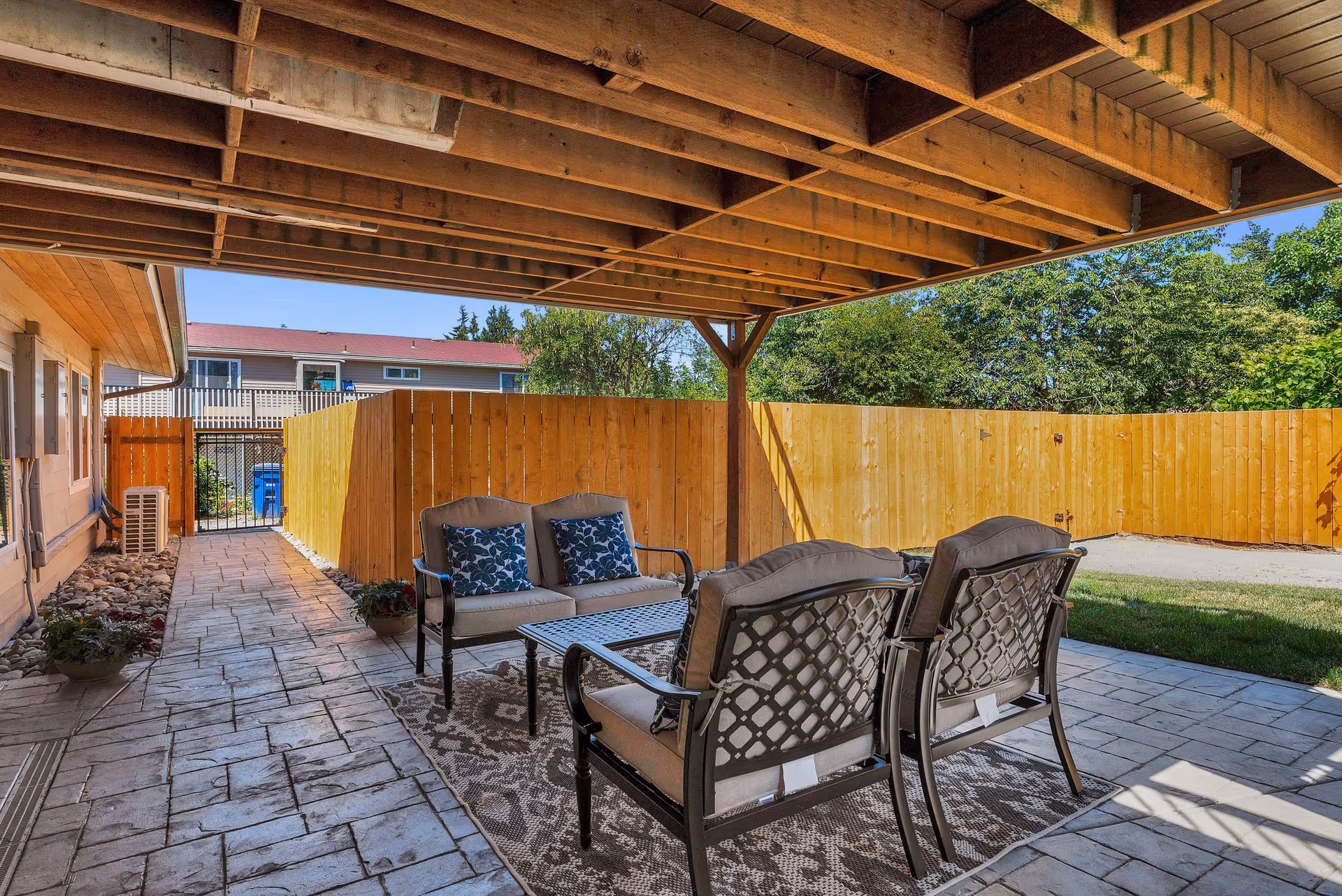 Covered outdoor patio area with a seating arrangement consisting of a loveseat and two chairs with cushions around a metal coffee table on a patterned rug. The patio has a wooden fence enclosing the space and a paved walkway leading to a gate. Trees and greenery are visible beyond the fence under a clear blue sky.