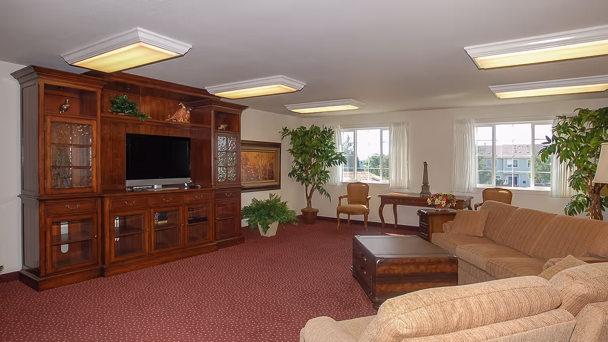 A spacious living room with a large wooden entertainment center holding a flat-screen TV, beige sectional sofa, wooden coffee table, two armchairs, side tables with decorative items, potted plants, and windows with white curtains letting in natural light.