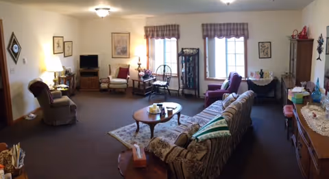 Spacious furnished living room with a patterned sofa, armchairs, coffee table, TV, and side tables beneath two windows.