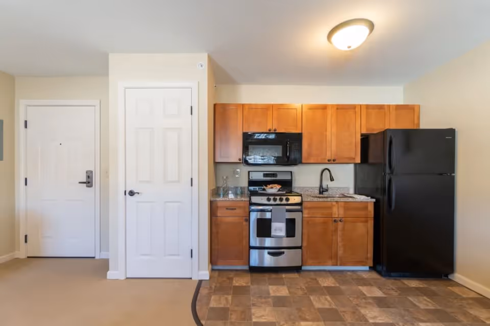 A small kitchen area with wooden cabinets, a black refrigerator, a stainless steel stove with oven, a black microwave above the stove, and a sink with a black faucet. To the left of the kitchen are two white doors, one likely an entrance door and the other a closet or pantry. The floor in front of the kitchen is tiled, while the rest of the floor is carpeted. The walls are painted light beige and there is a ceiling light fixture.