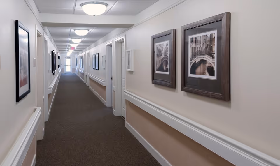 Long carpeted interior hallway with framed artwork, handrails, and ceiling lights in a senior living facility.