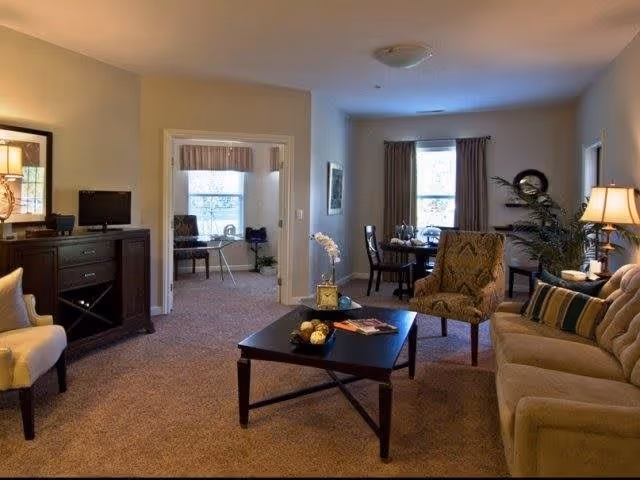 A cozy living room with beige carpet and walls, featuring a beige sofa with striped pillows, a patterned armchair, a dark wooden coffee table with decorative items, and a side table with a lamp. In the background, there is a dining area with a round table and chairs near a window with curtains, and an adjacent room with a desk and chair visible through an open doorway.