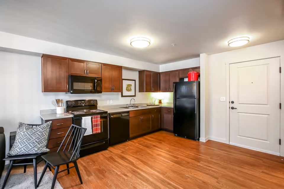 A modern kitchen area with dark wood cabinets, black appliances including a refrigerator, stove, microwave, and dishwasher. The kitchen has a light-colored countertop and a wooden floor. There is a black chair with a patterned cushion and a white door on the right side of the image.