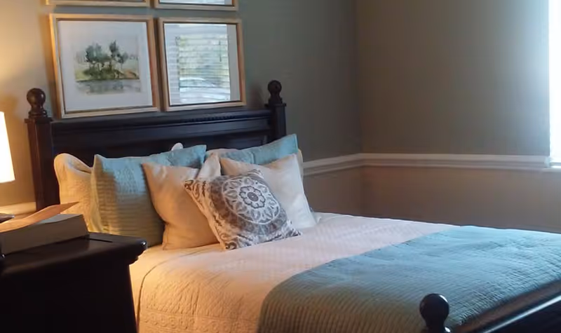A neatly made bed with a dark wooden headboard, adorned with multiple pillows including a decorative one with a circular pattern. The bed has a white quilt and a light blue throw blanket. There are framed pictures hanging on the wall above the headboard and a window letting in natural light on the right side.