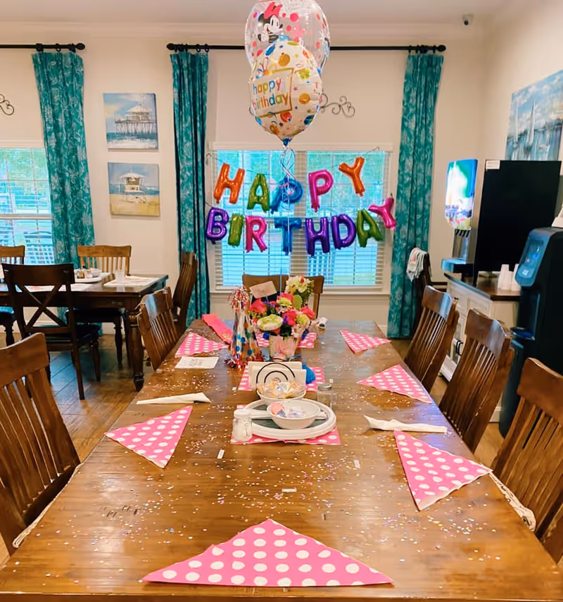 A dining room decorated for a birthday celebration with colorful 'HAPPY BIRTHDAY' balloons hanging in front of a window with teal curtains. A long wooden table is set with pink polka dot napkins, plates, bowls, and party hats, with confetti scattered across the table. There are wooden chairs around the table and additional tables and chairs in the background. The room has light-colored walls with beach-themed paintings and a beverage dispenser on a sideboard.