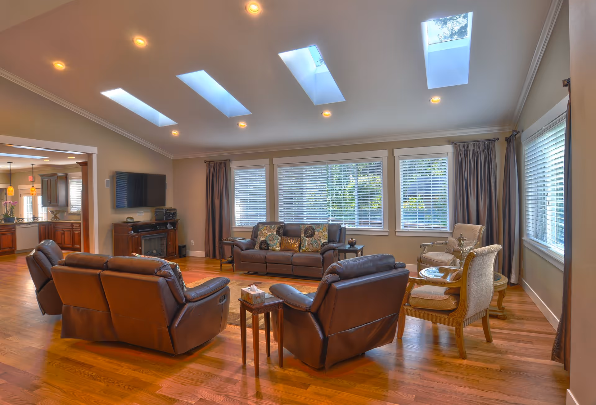 A spacious living room with wooden flooring, featuring a set of brown leather recliners and a matching sofa with decorative pillows. There are two upholstered armchairs with a small round table between them near the windows. The room has large windows with blinds and curtains, and skylights in the ceiling allowing natural light to brighten the space. A flat-screen TV is mounted on the wall above a wooden cabinet with a fireplace insert. The living room opens into a kitchen area visible in the background.