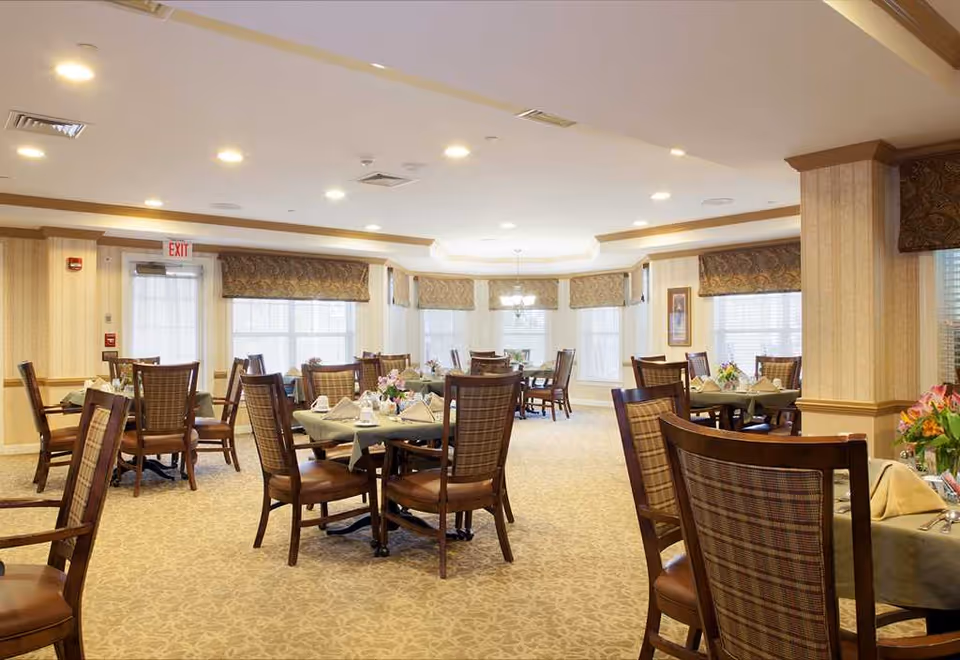 A bright and spacious dining room with multiple round tables set with tablecloths, napkins, and floral centerpieces. The room features large windows with patterned valances, beige walls with wooden trim, and carpeted floors. Several wooden chairs with cushioned seats and backs are arranged around the tables. Ceiling lights illuminate the space evenly.