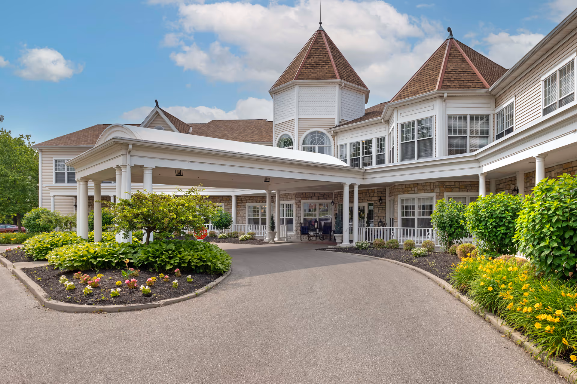 Exterior view of Brookdale Kenwood senior living facility featuring a covered entrance with white columns, landscaped flower beds, and a paved driveway. The building has multiple windows and two turret-style roof structures under a partly cloudy sky.