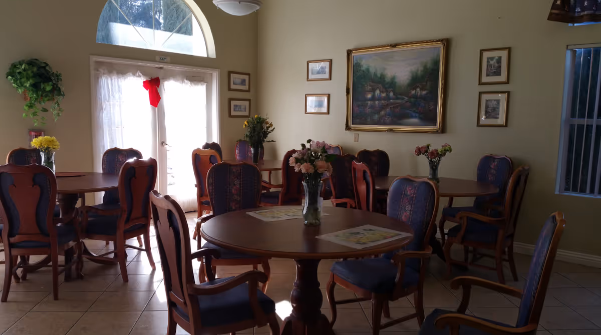 Sunlit communal dining room with round wooden tables, upholstered chairs, and floral centerpieces.