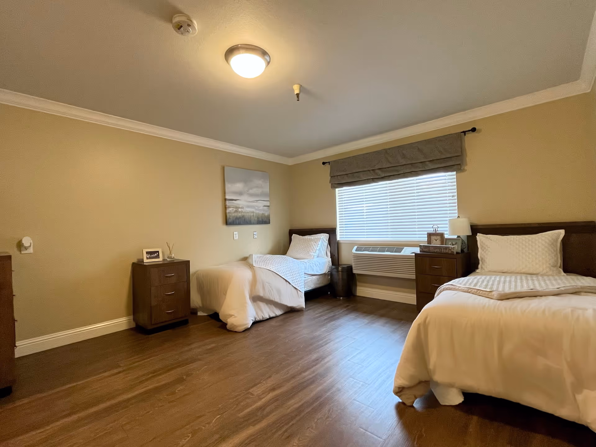 A bedroom with two single beds, each with white bedding and pillows. Between the beds are two wooden nightstands with decorative items including a lamp, a clock, and framed pictures. A window with closed blinds and a fabric valance is centered on the wall behind the beds. The room has beige walls, wooden flooring, and a ceiling light fixture.
