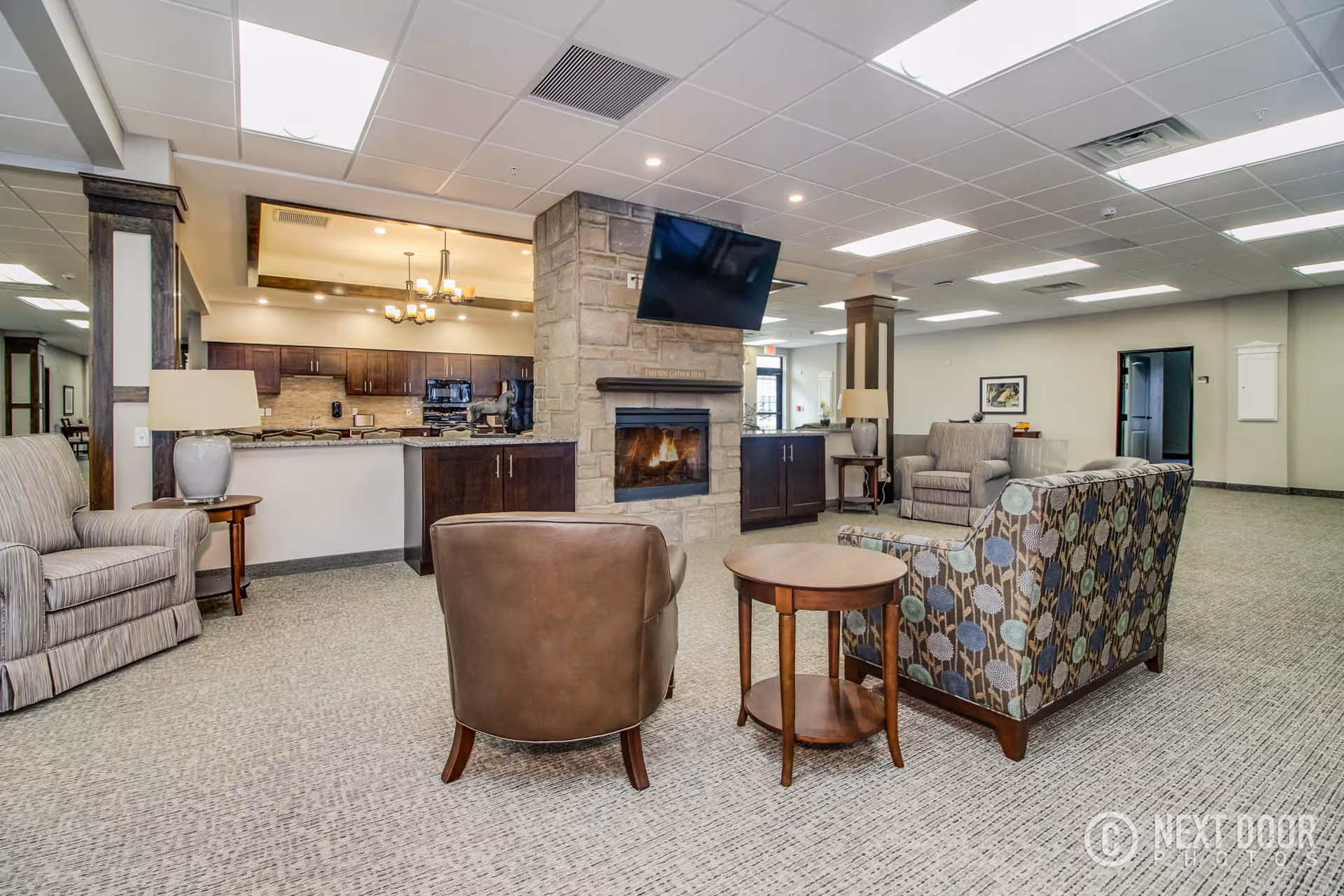 Community lounge with armchairs and a patterned sofa arranged around a stone fireplace and wall-mounted TV, with a kitchenette visible in the background.