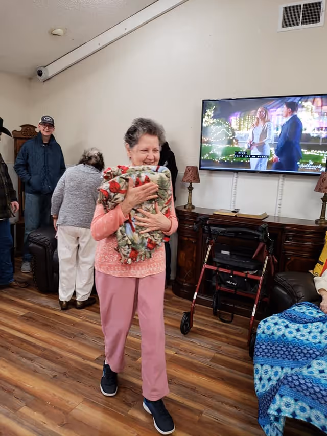 An elderly woman smiling and holding a colorful blanket in a room with wooden flooring. In the background, several other elderly people are present, some sitting and some standing. A flat-screen TV mounted on the wall shows a scene from a Hallmark movie. There is a wooden cabinet beneath the TV with a walker placed in front of it, and two small lamps on either side of the cabinet.