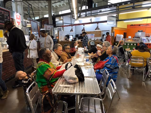 A group of elderly people sitting and eating at a long metal table in a busy indoor dining area with other patrons and food service counters in the background.
