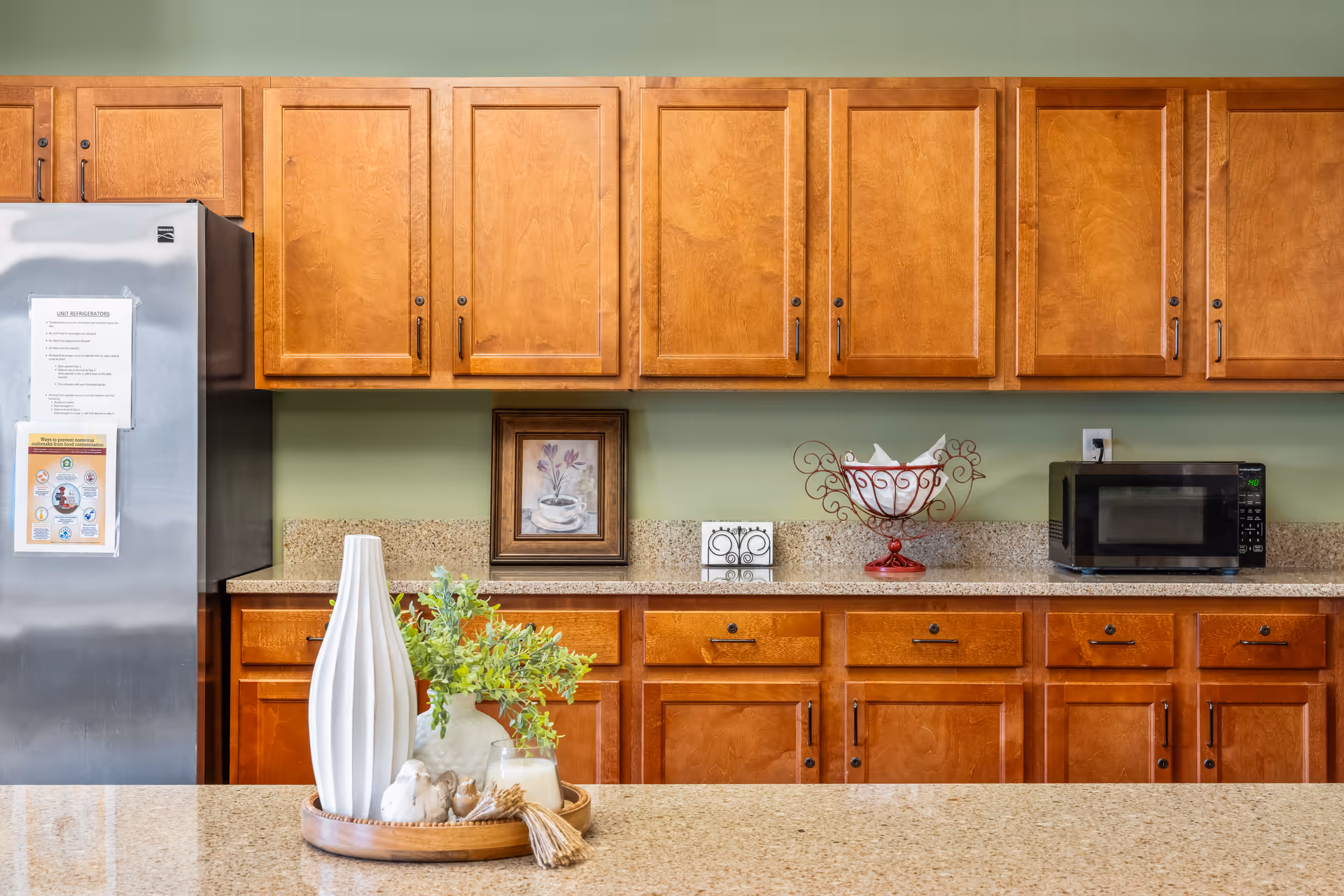 A kitchen area with wooden cabinets and drawers, a stainless steel refrigerator on the left, a microwave on the right, and a granite countertop. On the countertop in the foreground, there is a decorative tray with a white vase, greenery, and small ornamental items. A framed picture and a decorative tissue holder are on the back counter against a light green wall.