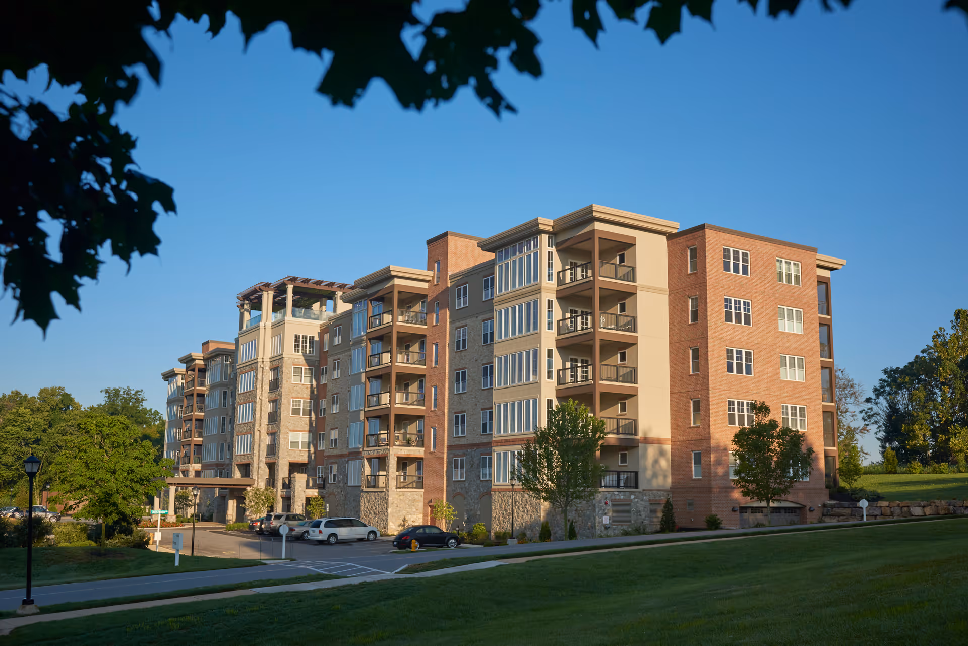 Exterior view of a multi-story senior living facility building with balconies, surrounded by green lawns and trees under a clear blue sky.