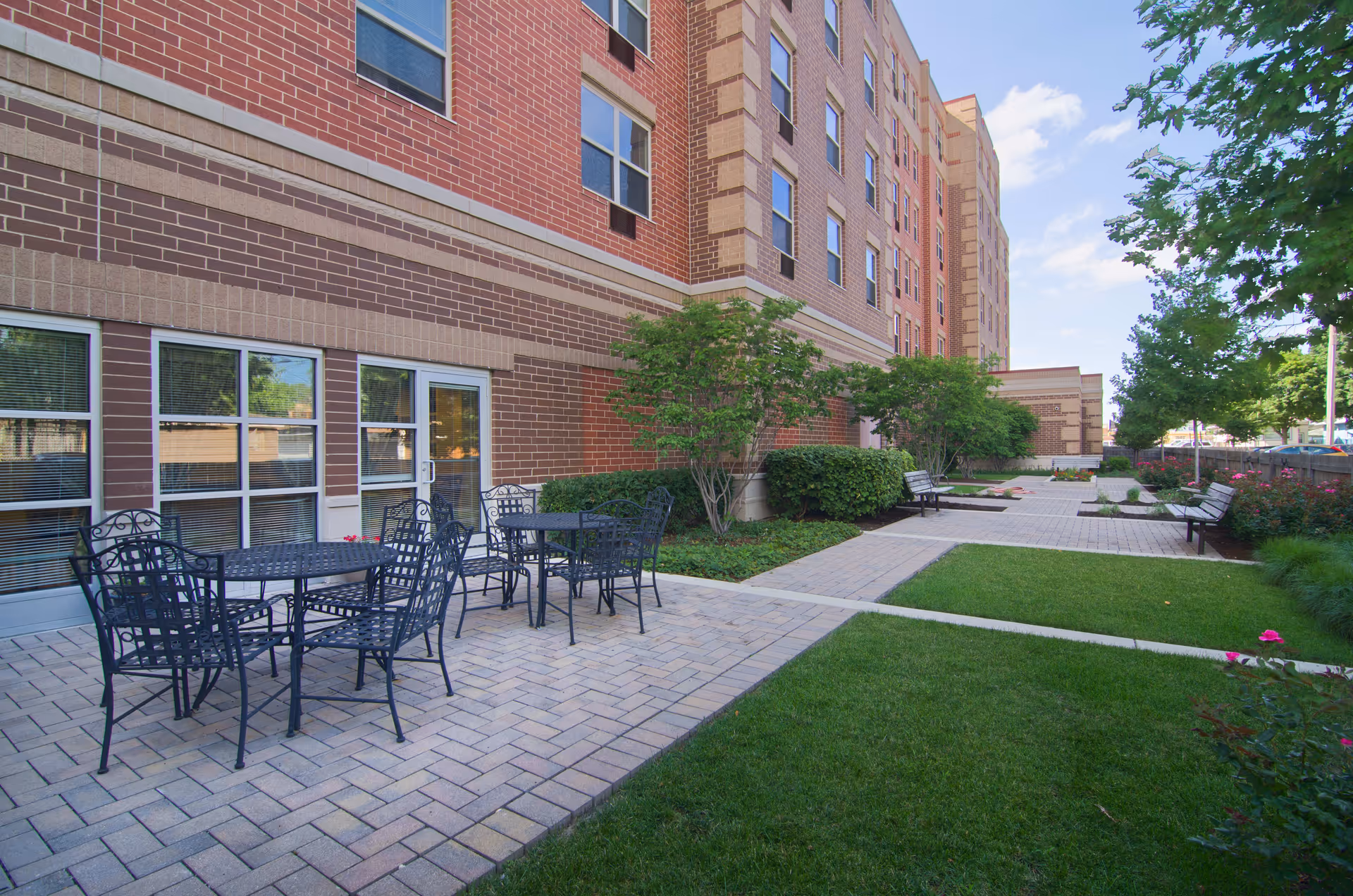 Outdoor patio area at Senior Suites of Kelvyn Park with black metal tables and chairs on a paved surface next to a brick building. There are green bushes, trees, and a well-maintained lawn surrounding the patio, with benches along the walkway.