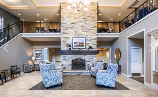 Two-story lobby/lounge with a central stone fireplace, patterned armchairs arranged on a rug, and an upper balcony overlooking the space.