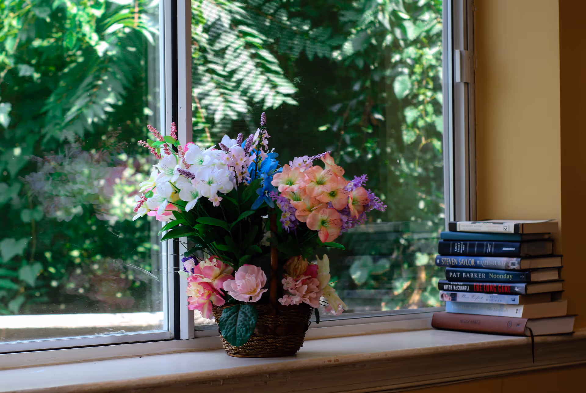 A window sill with a basket of colorful artificial flowers and a stack of books next to it. Outside the window, green foliage is visible.