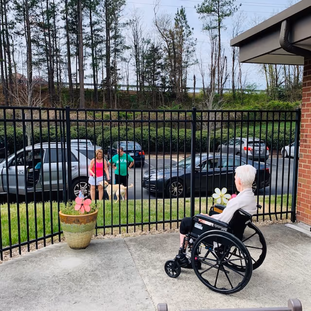 An elderly person in a wheelchair on a concrete patio area near a black metal fence, looking at two people standing outside the fence with a dog. There are several parked cars and trees in the background.