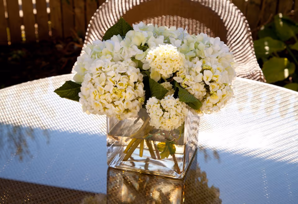 A glass vase of white hydrangea flowers on a reflective outdoor glass table with a wicker chair in the background.