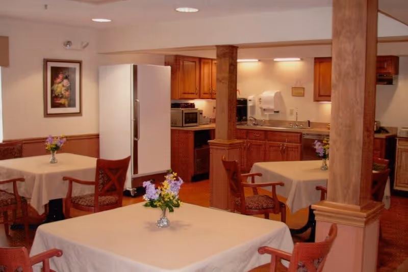 Dining area with tables draped in white cloths and small flower vases, wooden chairs, and a kitchenette with refrigerator and cabinets in the background.