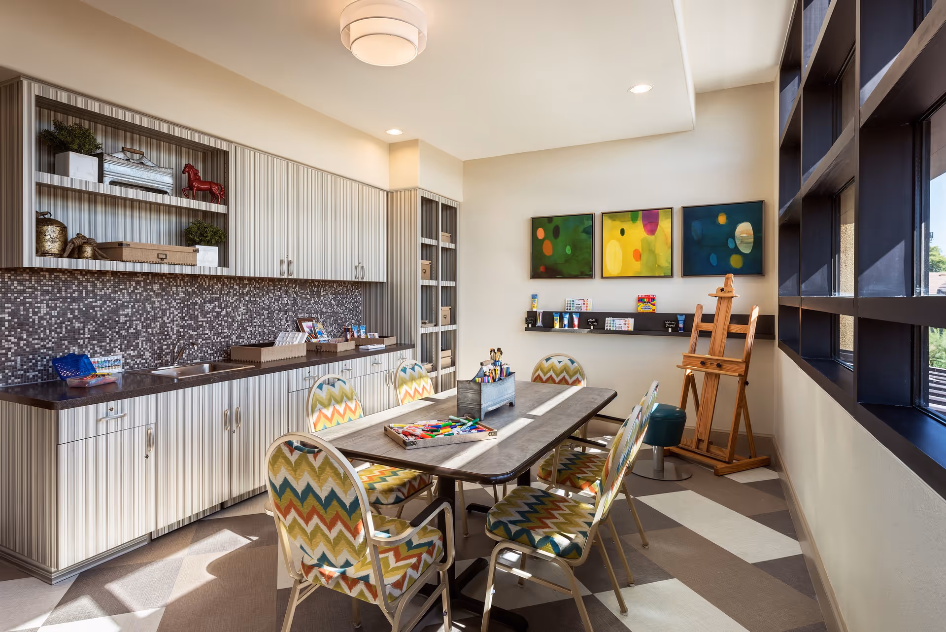 A bright and colorful art room with a rectangular table surrounded by six patterned chairs. The room features a countertop with a sink and cabinets above and below, decorated with plants and art supplies. On the wall, three abstract paintings hang above a shelf holding various art materials. An easel and a stool are positioned near large windows letting in natural light.