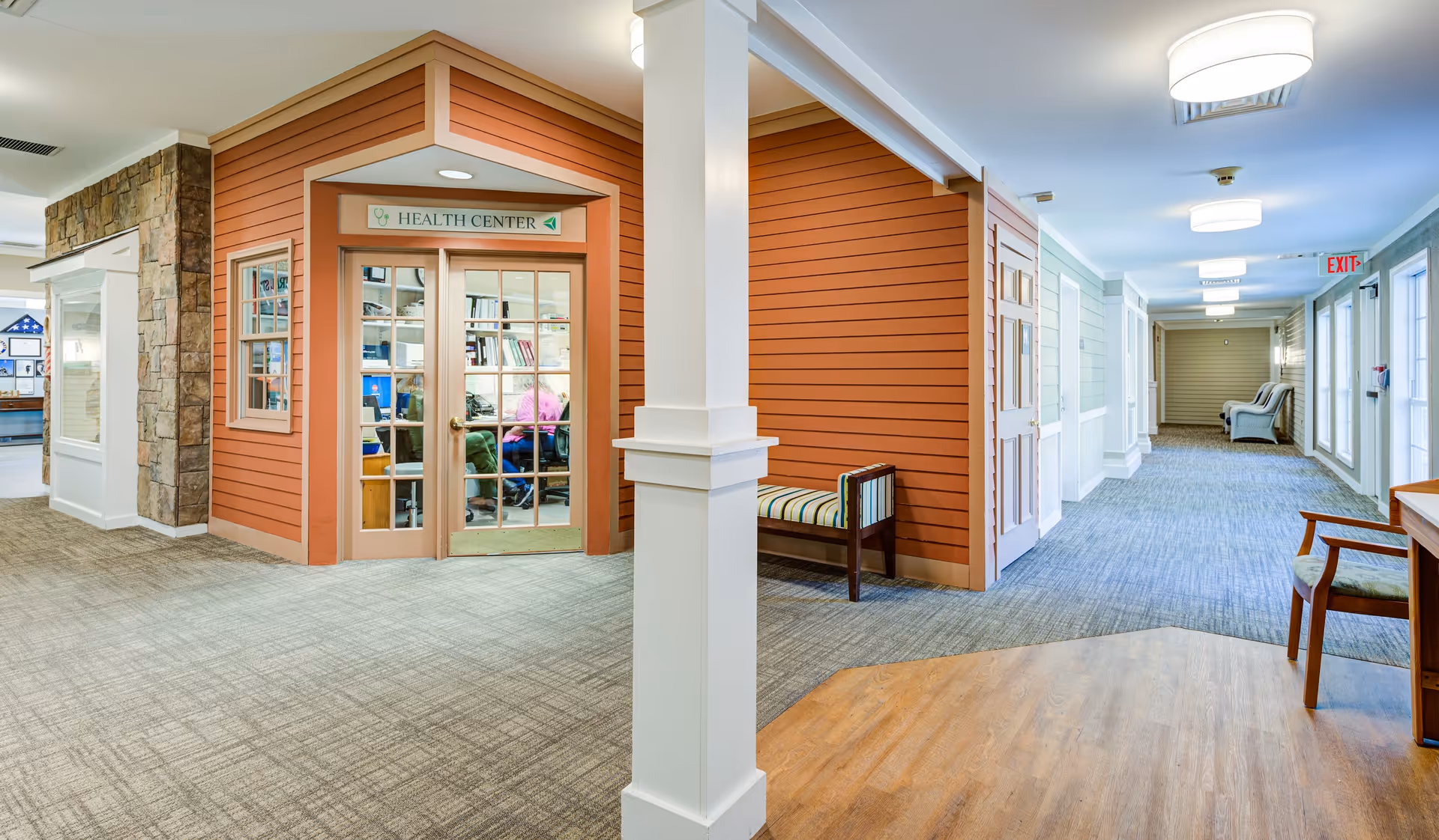 Interior hallway of a senior living facility featuring a health center with glass double doors and a sign above. The hallway has carpeted floors, a wooden bench with striped upholstery, and several chairs along the walls. The walls are painted in warm tones with white trim, and there are ceiling lights providing bright illumination.