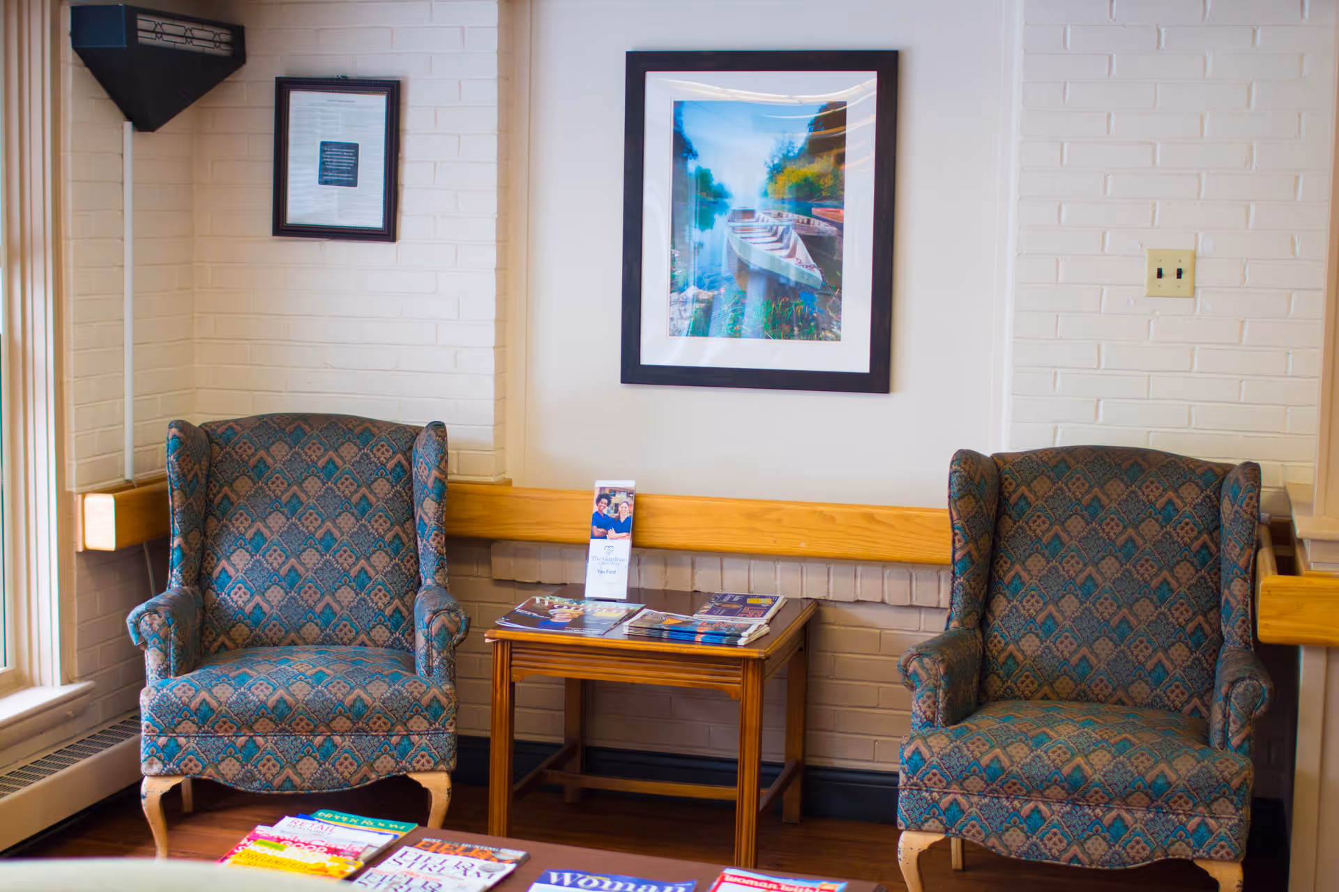 Two patterned armchairs flank a small wooden table with magazines beneath a framed waterside photo on a white brick wall in a waiting area.