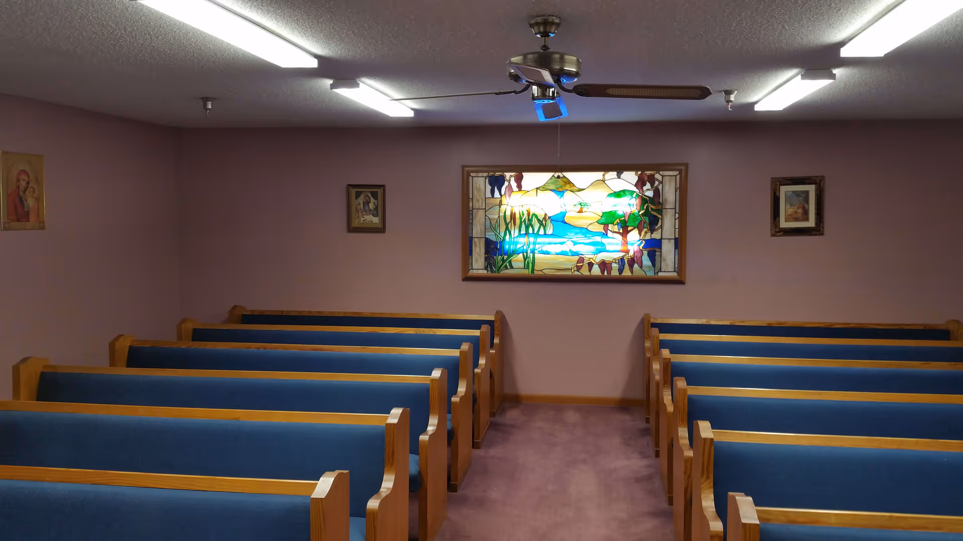 Interior view of a small chapel or prayer room with wooden pews upholstered in blue fabric arranged in rows facing a wall with a colorful stained glass window depicting a nature scene. The walls are painted light purple and decorated with framed religious artwork. Ceiling lights and a ceiling fan are visible above.