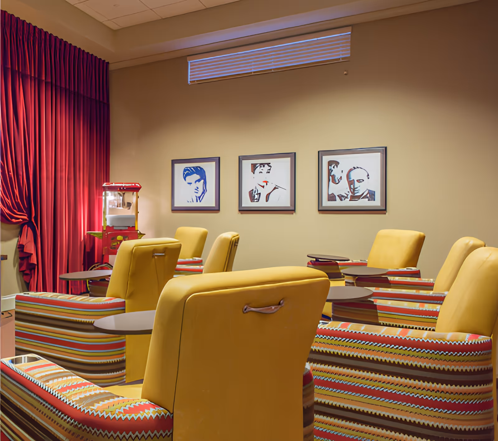 A cozy lounge area with several yellow and striped armchairs arranged in rows facing a wall with three framed black and white portraits. A red popcorn machine is positioned near a window with red curtains.