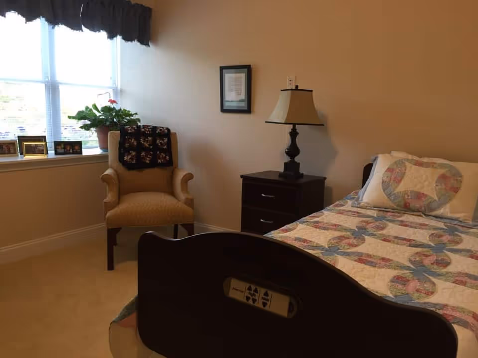 A cozy bedroom with a single bed covered in a colorful quilt and matching pillow. Next to the bed is a dark wooden nightstand with a lamp on it. A beige upholstered armchair with a dark patterned throw draped over the back is positioned near a window with blinds and a dark valance. On the windowsill are several framed photos and a potted plant.