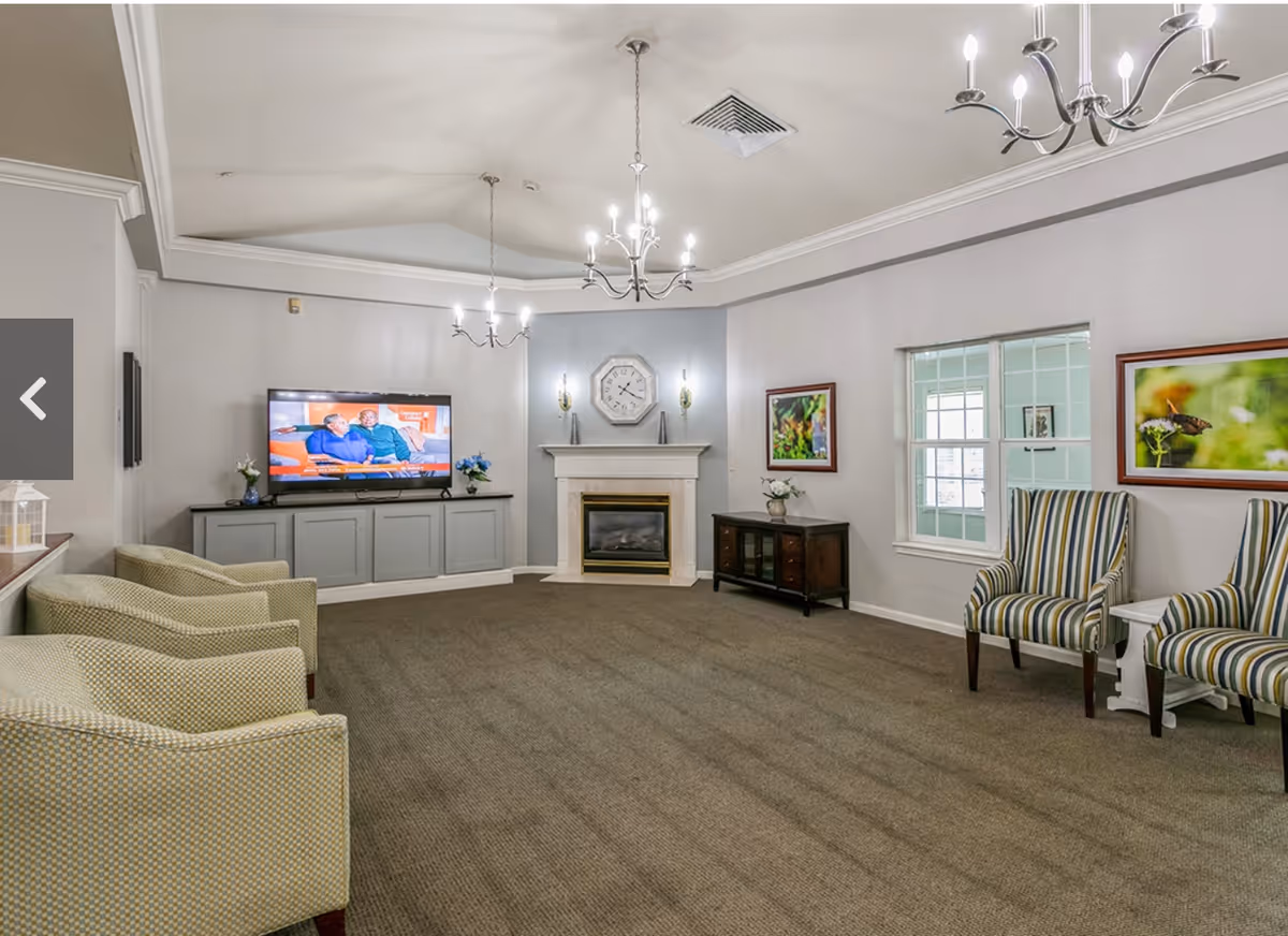 A spacious living room with a beige carpet, multiple armchairs arranged along the walls, a flat-screen TV on a gray cabinet, a white fireplace with a clock above it, two chandeliers hanging from the ceiling, and framed nature photographs on the walls.
