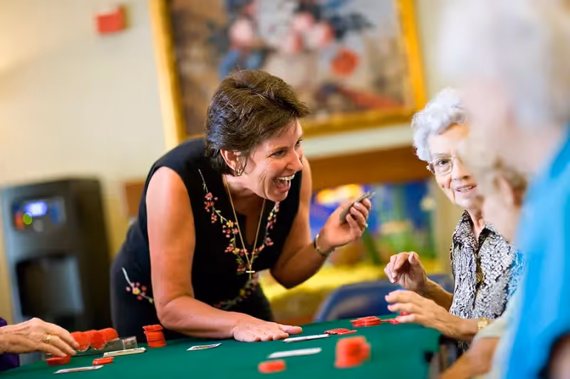A group of elderly people and a woman are gathered around a green poker table playing a card game. The woman is smiling and holding cards, engaging enthusiastically with the group. The background shows a blurred painting and some furniture.
