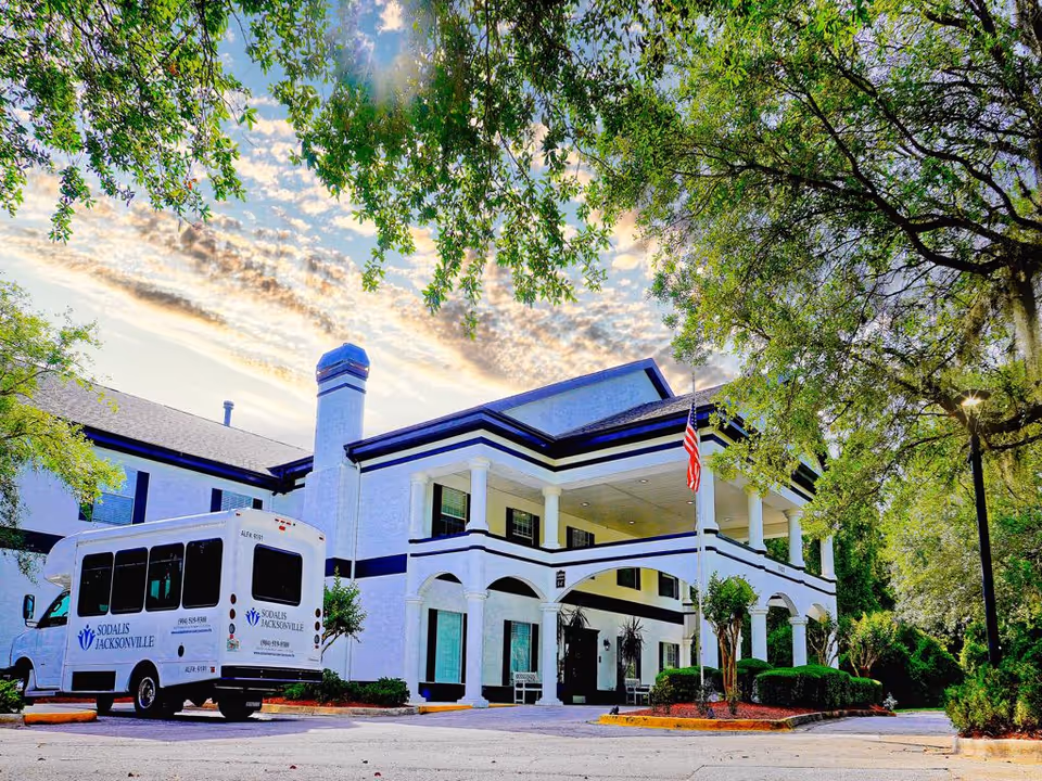 Exterior view of Sodalis Jacksonville facility showing a two-story white building with black trim, a covered entrance with columns, an American flag, and a white shuttle bus parked in front. The scene is framed by green trees under a partly cloudy sky.
