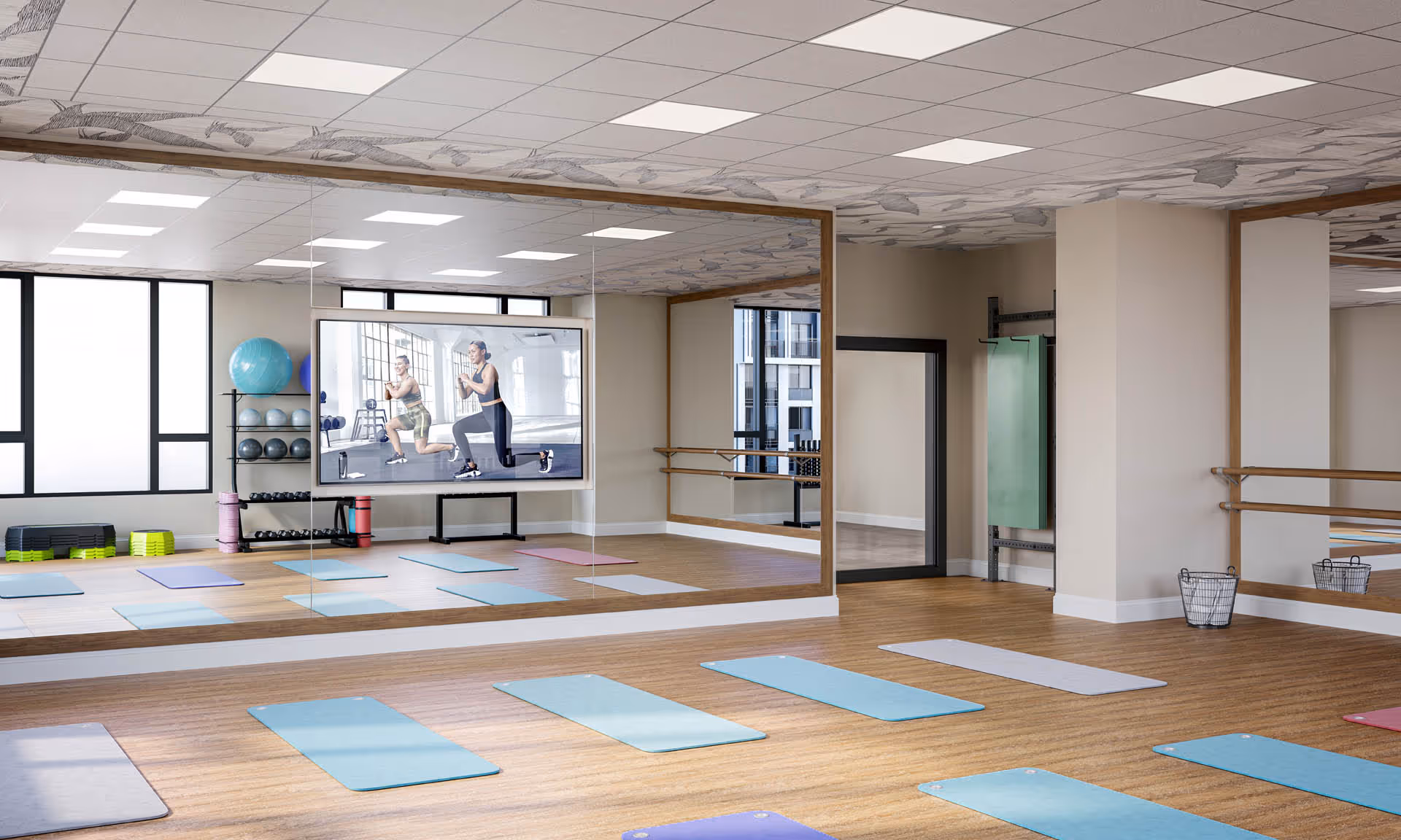 A spacious exercise room with wooden flooring and large mirrors on the walls. Several colorful yoga mats are laid out on the floor. There is a large screen mounted on the mirrored wall displaying two people exercising. Shelves with exercise balls, weights, and other fitness equipment are visible near the windows.