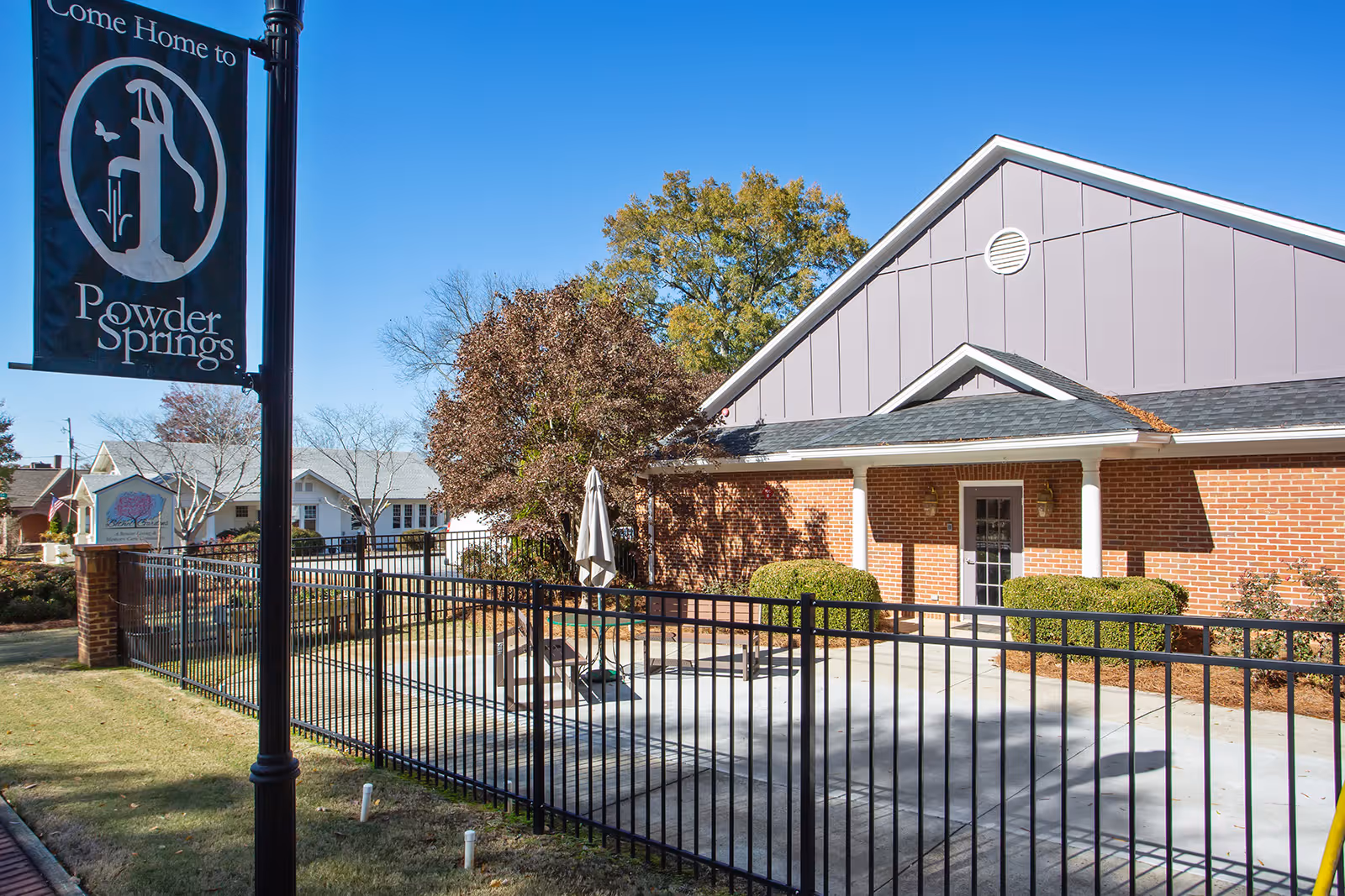 Brick assisted-living building with a fenced concrete courtyard, patio table and umbrella, landscaping, and a 'Powder Springs' street banner.