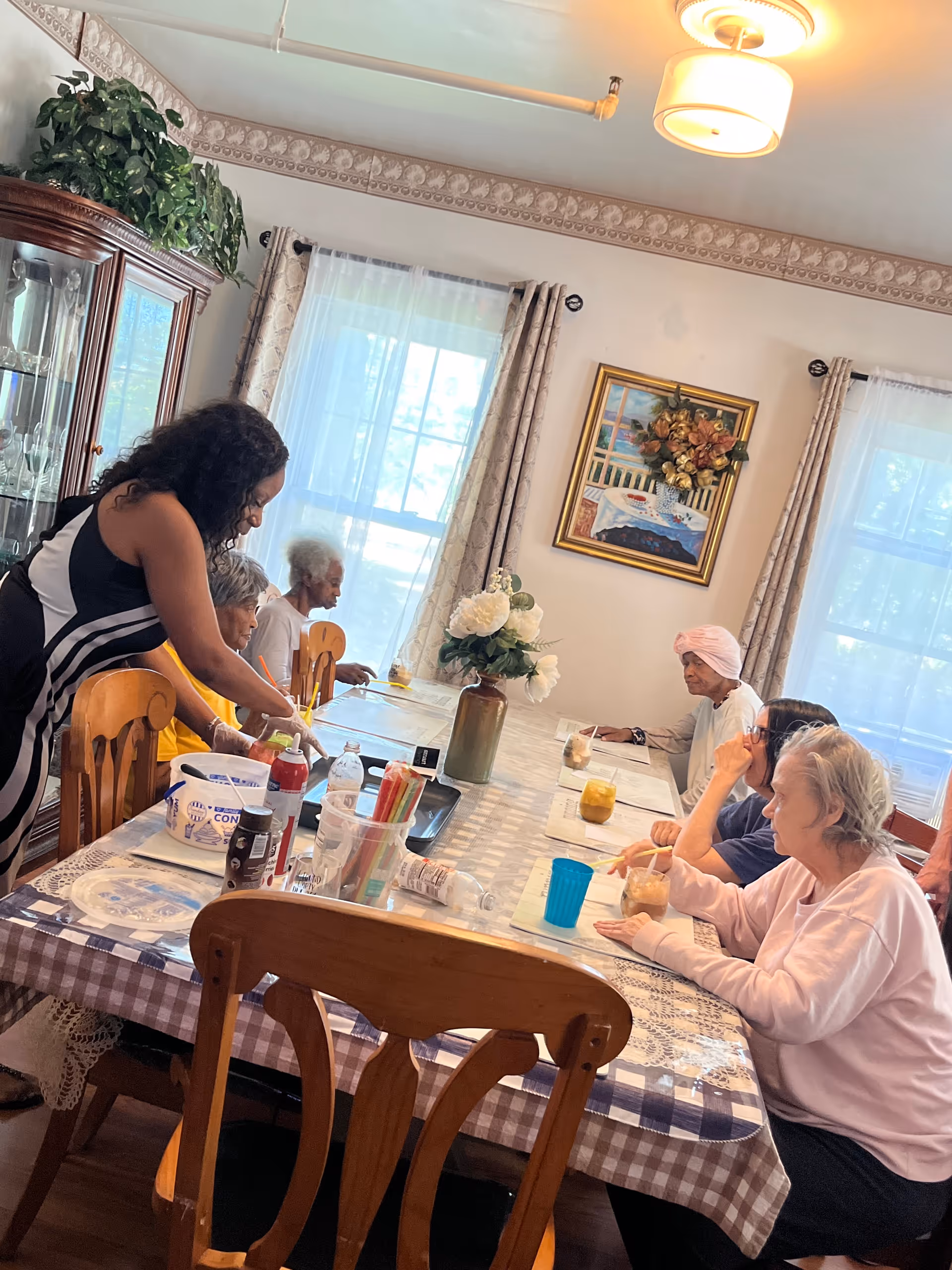 A group of elderly women sitting around a dining table in a well-lit room with large windows and curtains. A younger woman is standing and assisting one of the elderly women. The table is covered with a checkered tablecloth and has various items including drinks, art supplies, and a vase with white flowers. There is a painting on the wall and a wooden cabinet with glassware in the background.