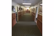 A long hallway in a healthcare facility with carpeted floor, beige walls with wooden paneling, framed artwork on the walls, and doors leading to rooms on both sides. The hallway is well-lit with ceiling lights and has some decorative plants placed along the walls.