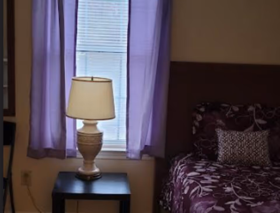 A bedroom with a purple bedspread, nightstand, lamp, and purple curtains.