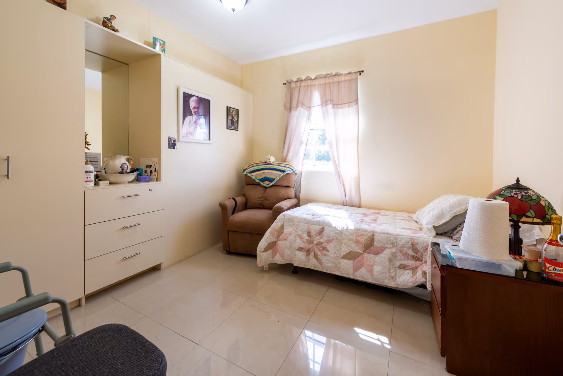 A small, cozy bedroom in an assisted living facility with a single bed covered in a patterned quilt, a brown armchair with a colorful blanket draped over the back, a window with sheer curtains letting in natural light, a wooden nightstand with a decorative lamp and various items, and a built-in cabinet with drawers and a mirror.