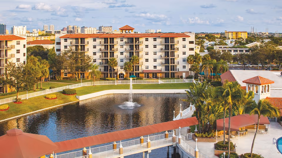 Exterior view of a multi-story residential building overlooking a pond with a central fountain, walkways, and palm trees.
