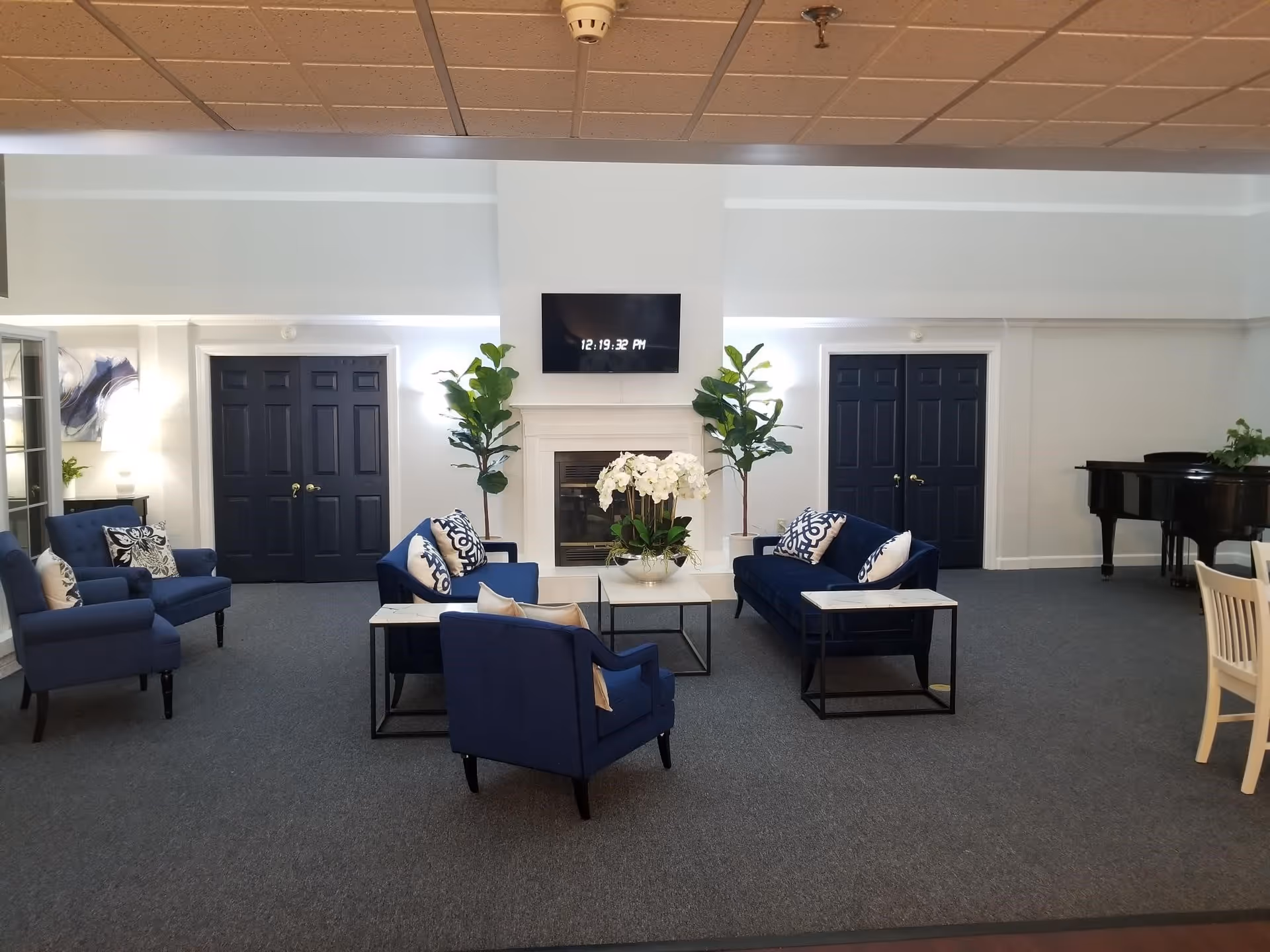 A spacious living room area with blue upholstered chairs and sofas arranged around a white coffee table with a flower arrangement. There are two tall green plants flanking a white fireplace with a mounted flat-screen TV above it displaying the time 12:19:32 PM. The room has dark double doors on either side of the fireplace, a grand piano in the right corner, and a dining table with chairs partially visible on the right side. The ceiling has a grid pattern with recessed lighting.