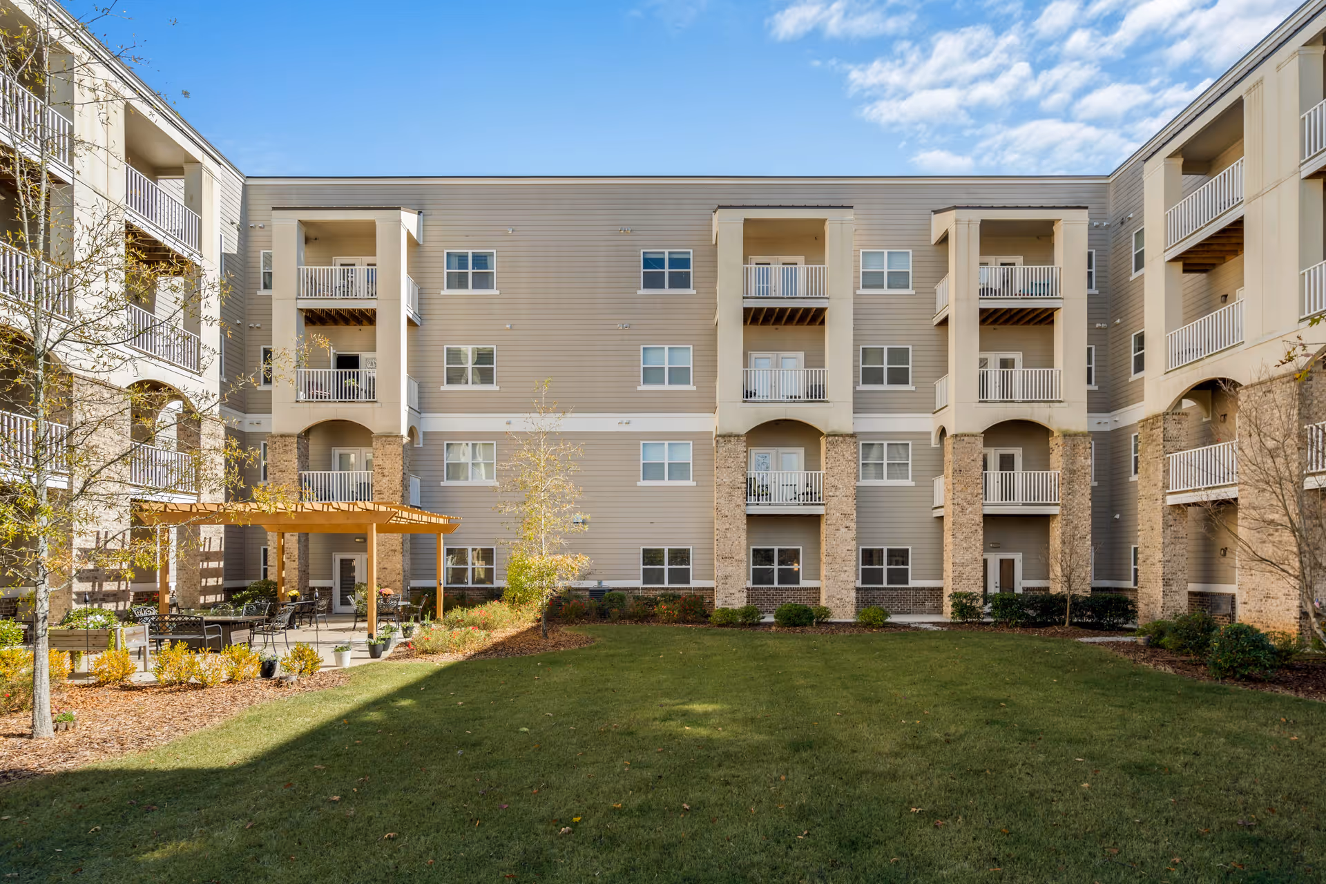Outdoor courtyard area of The Mansions at Alpharetta Independent Living featuring a well-maintained green lawn, a wooden pergola with seating underneath, surrounded by a multi-story building with balconies and windows under a partly cloudy blue sky.