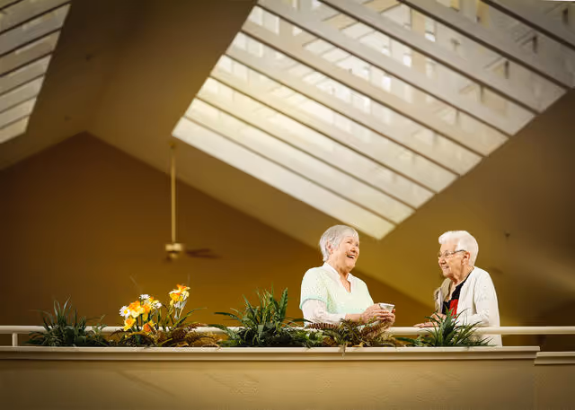 Two elderly women smiling and talking to each other while standing near a railing adorned with green plants and yellow flowers, under a large skylight ceiling with a ceiling fan in the background.