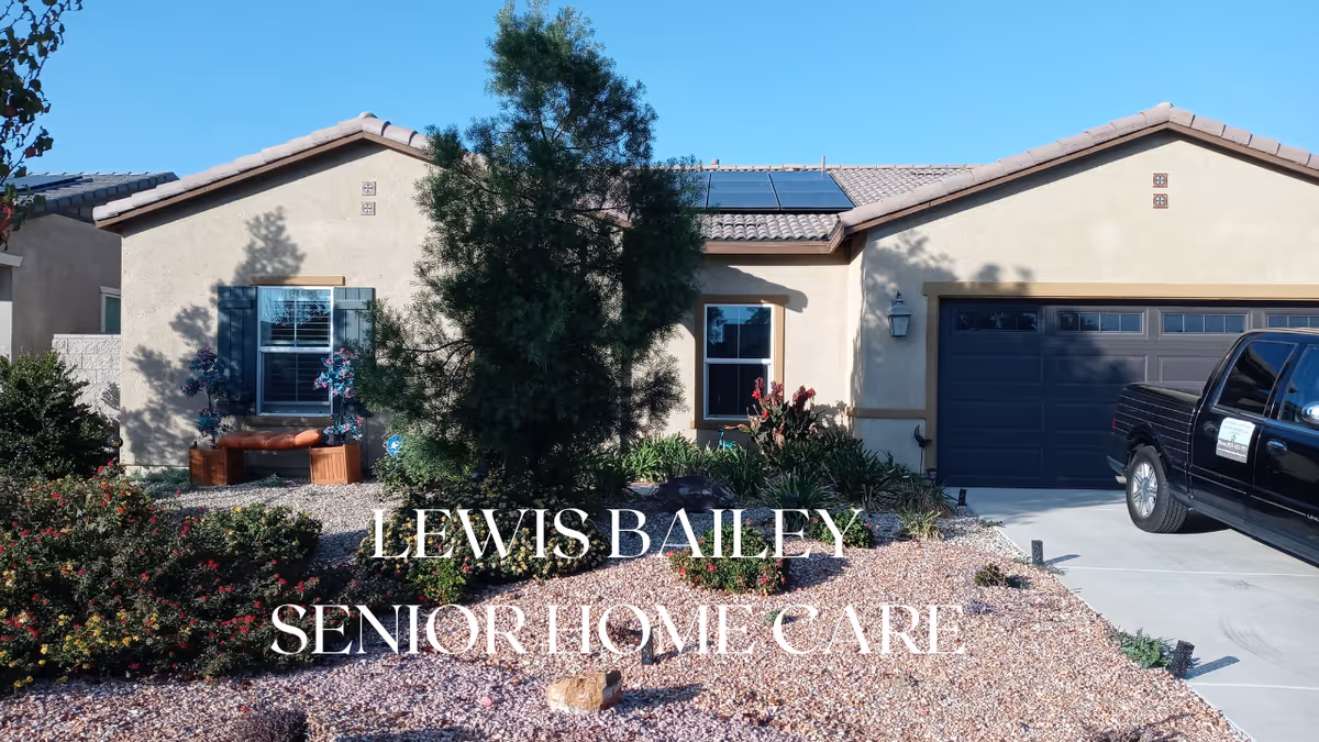 Exterior view of a single-story beige stucco house with a tiled roof, a small garden with bushes and a tree in front, a black pickup truck parked in the driveway, and clear blue sky above.