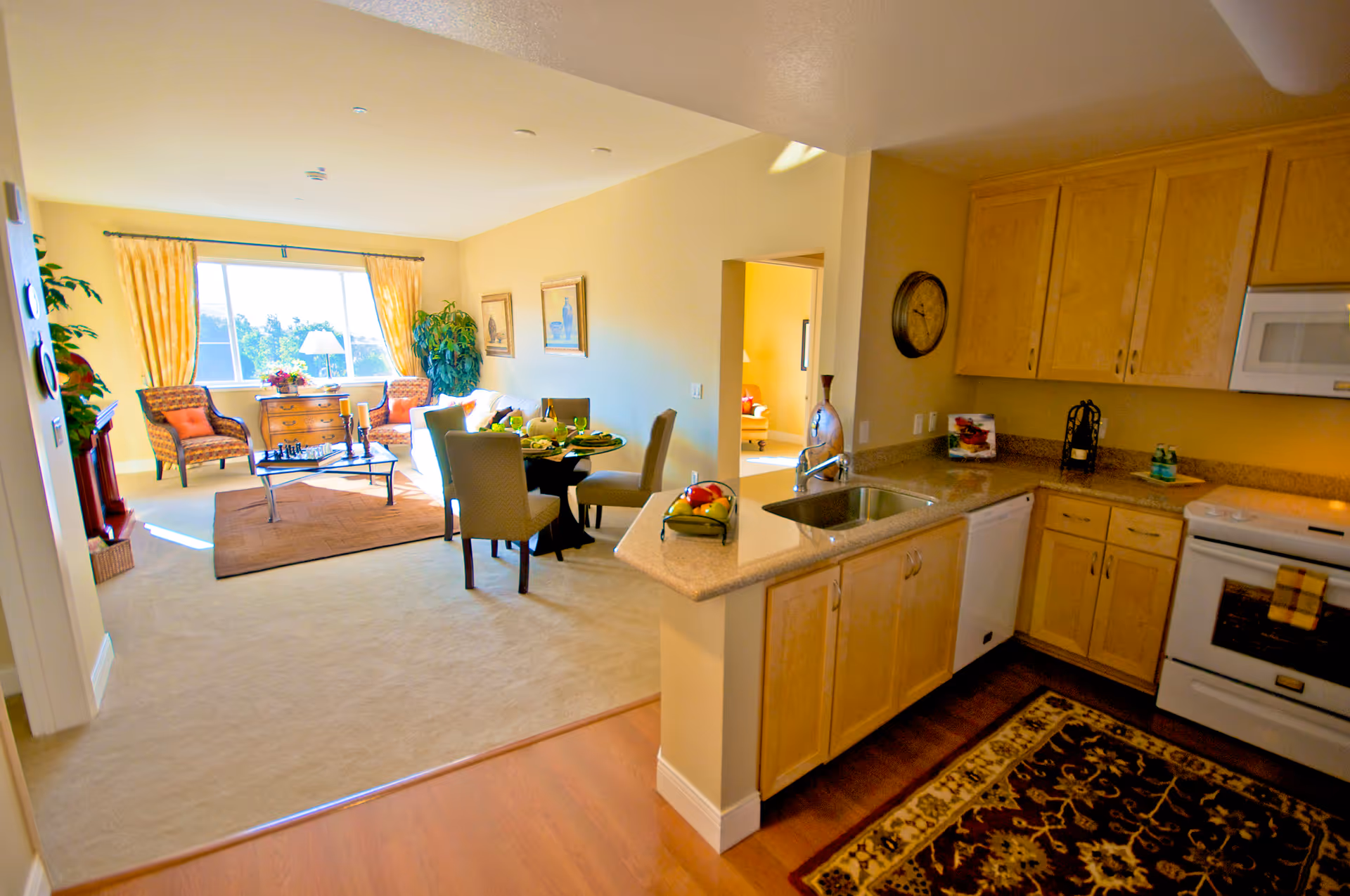 Interior view of a retirement community apartment showing a kitchen area with wooden cabinets, a sink, dishwasher, stove, and microwave. Adjacent to the kitchen is a dining area with a round table and four chairs, set with plates and glasses. Beyond the dining area is a living room with two armchairs, a sofa, a coffee table with candles and a chessboard, a chest of drawers, and large windows with yellow curtains letting in natural light.
