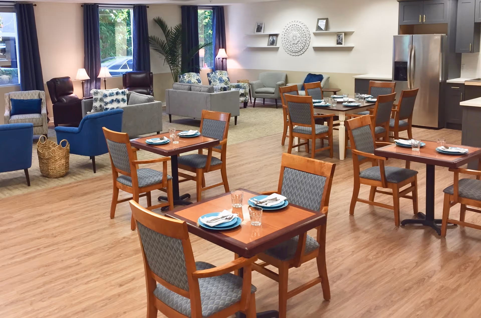 Spacious senior living dining area with multiple set wooden tables and chairs and a comfortable lounge with sofas and armchairs in the background.