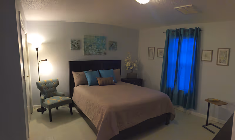 A cozy bedroom with a bed, nightstands, teal curtains, an upholstered chair, and wall art.
